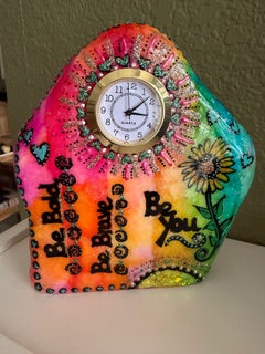Colorful painted stone with a clock and decorative elements on a plain background. Text painted on the stone "Be Bold, Be Brave, Be You."