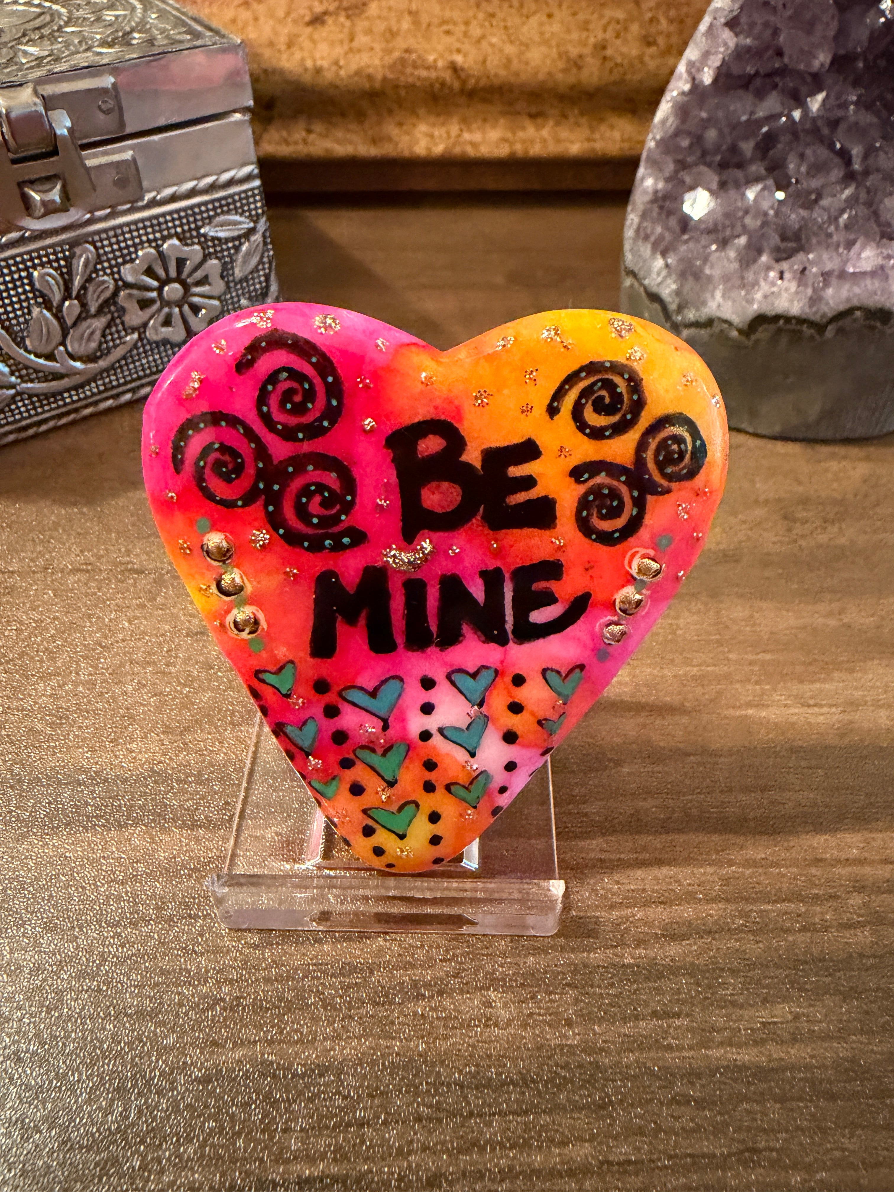 Heart-shaped painted stone with 'Be Mine' text on a textured surface