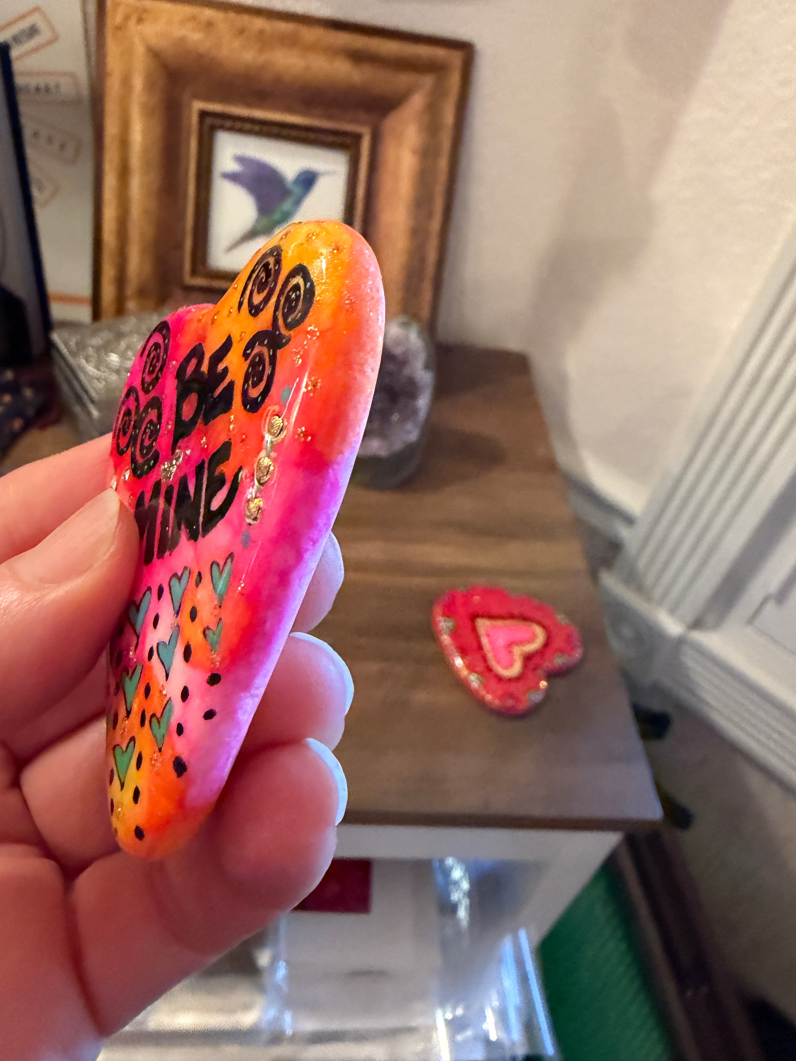 Colorful painted heart with decorative patterns held by a hand in an indoor setting.