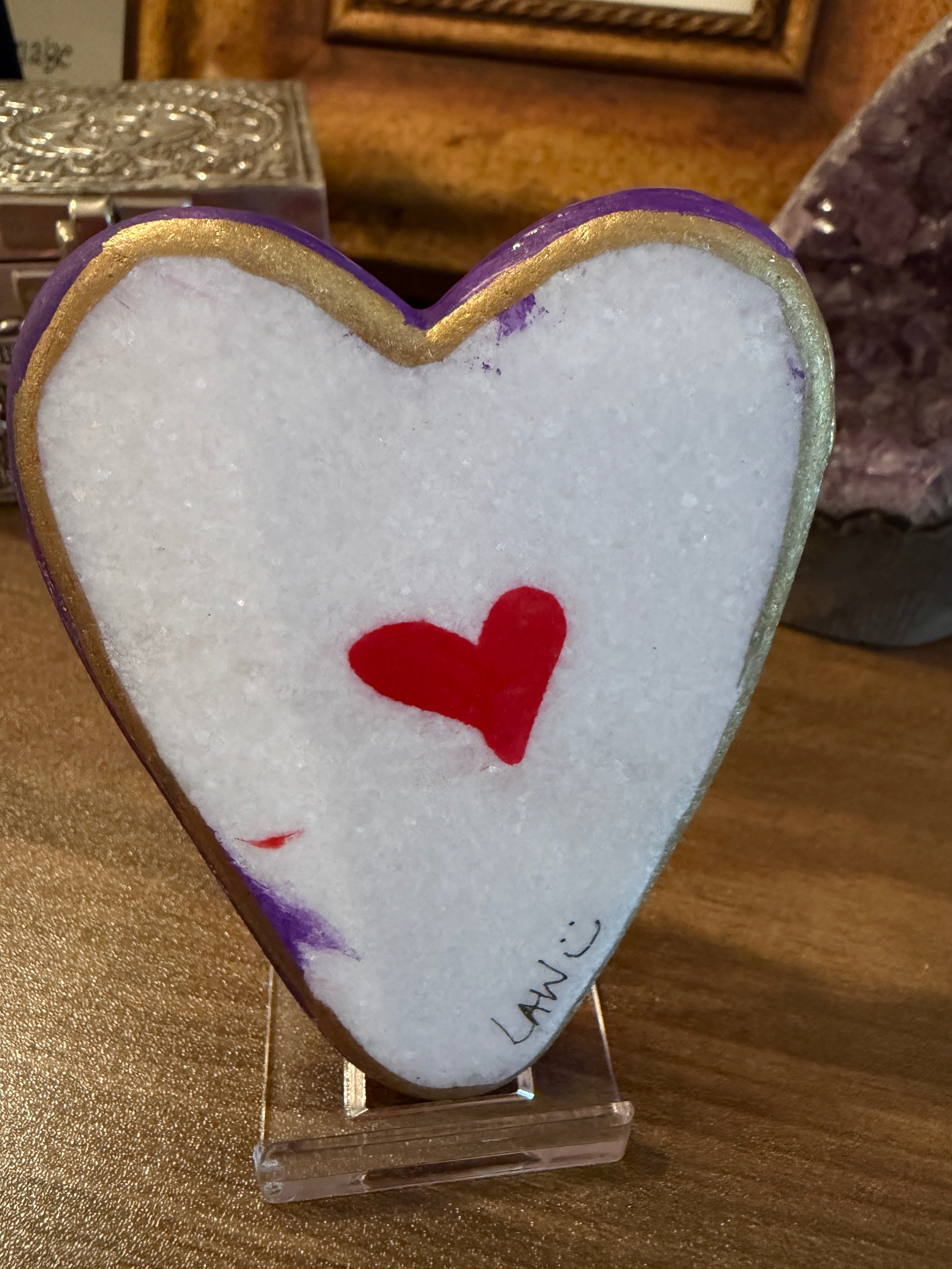 Heart-shaped painted stone with a red heart design on a white background, placed on a wooden surface.