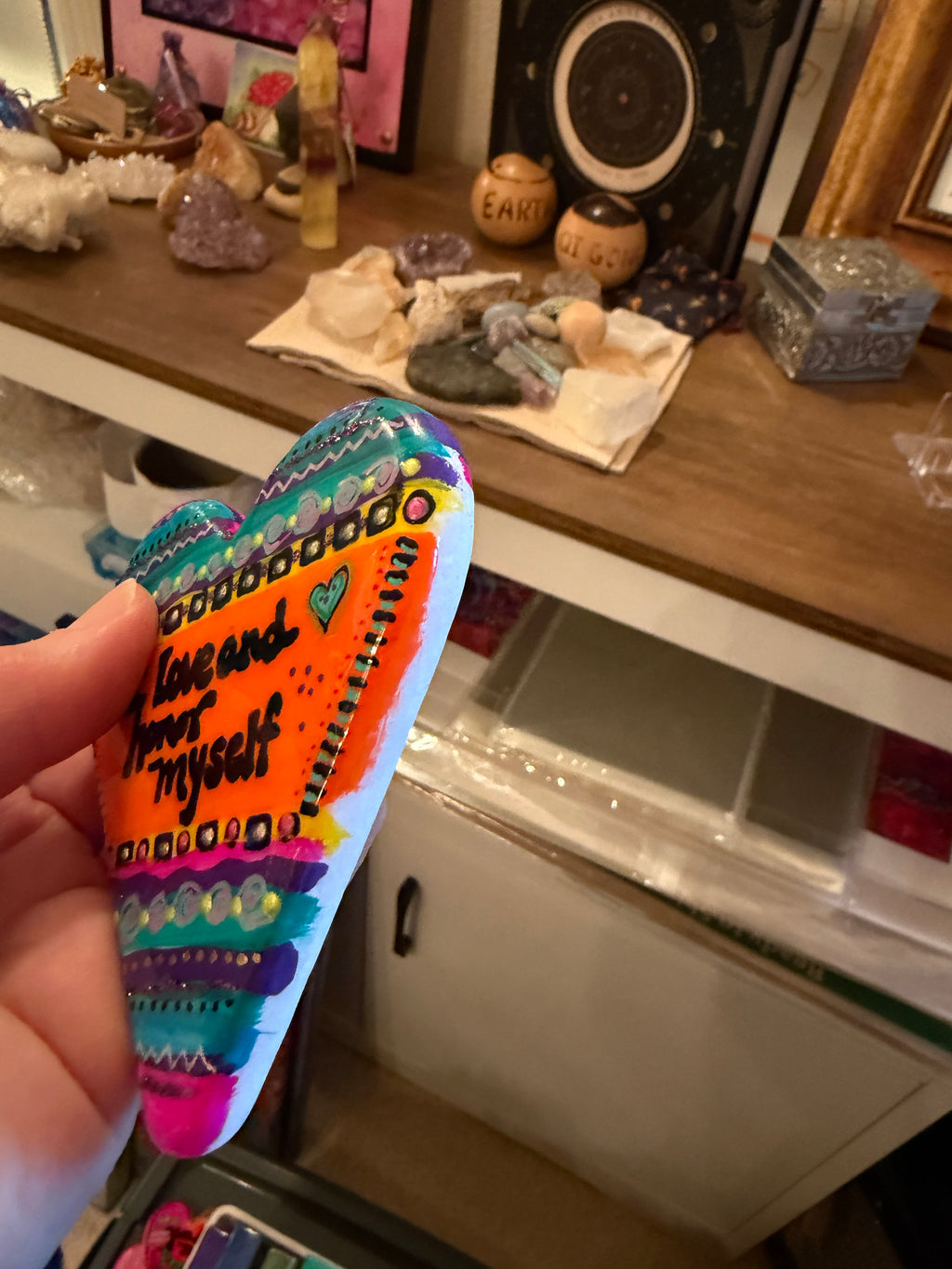 Colorful painted heart stone with text held in front of a shelf with decorative items