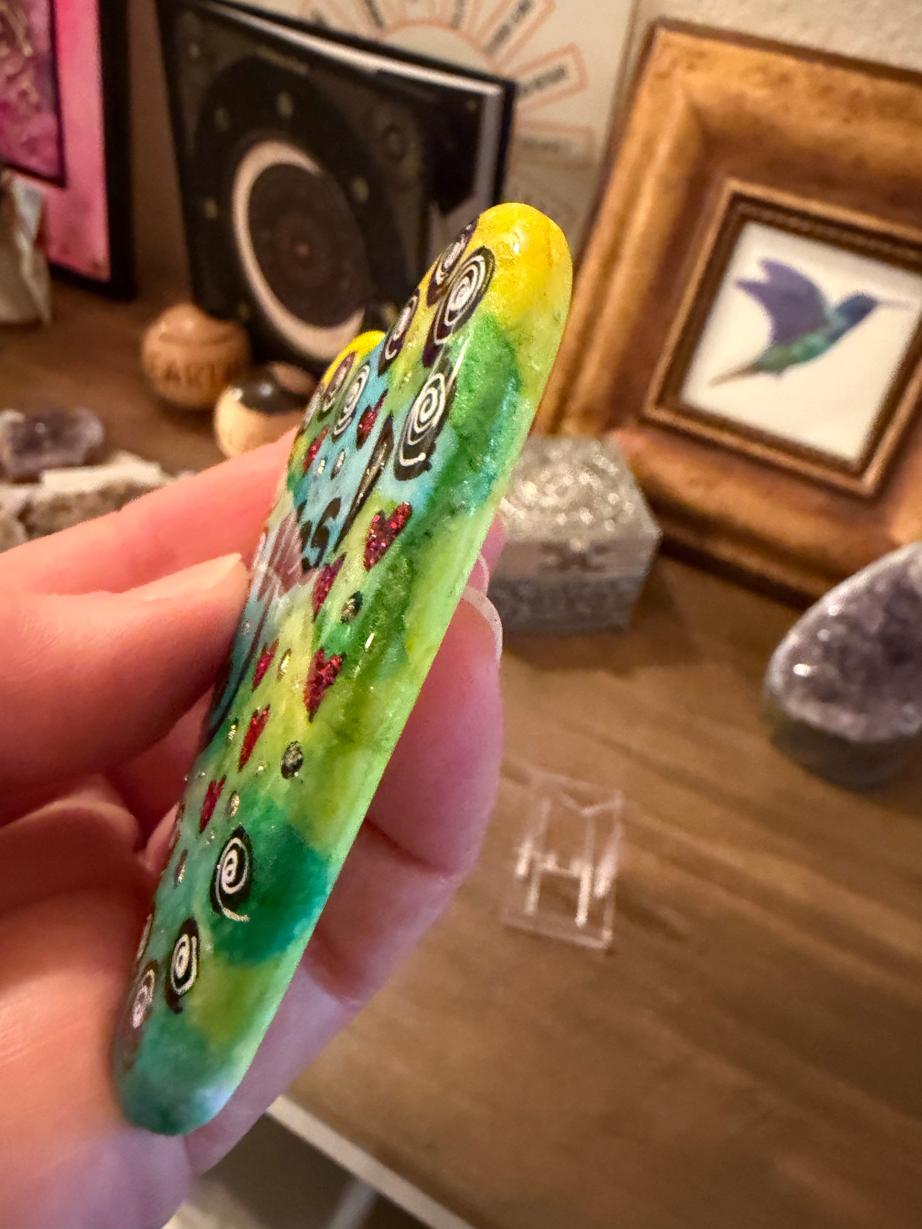 Hand holding a painted heart stone with decorative patterns against a blurred indoor background
