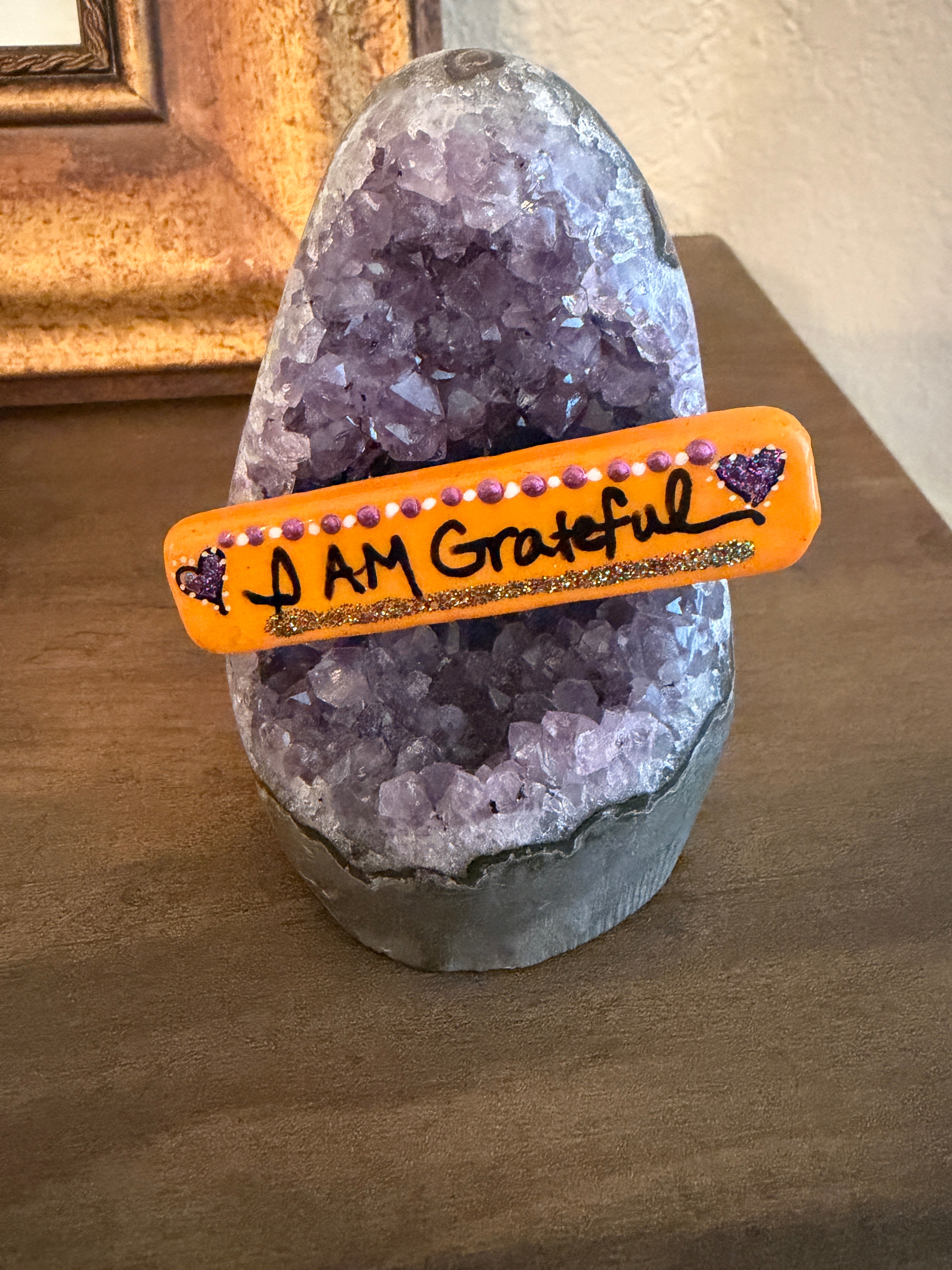 A handpainted stone stick reading 'I Am Grateful' on an amethyst stand.