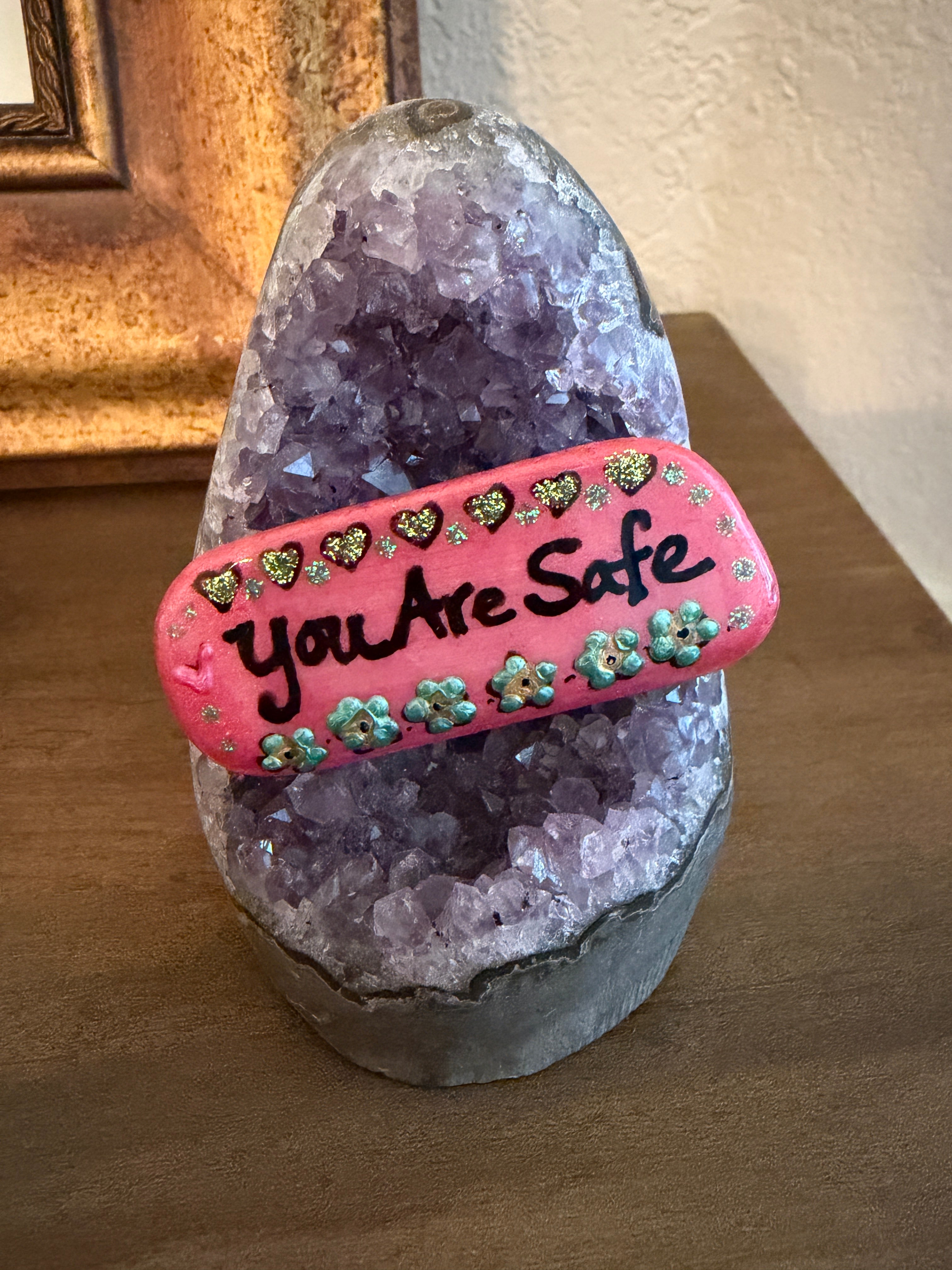 Pink decorative stone with an affirmation 'You Are Safe' painted on it. Stone is resting on an amethyst stand.
