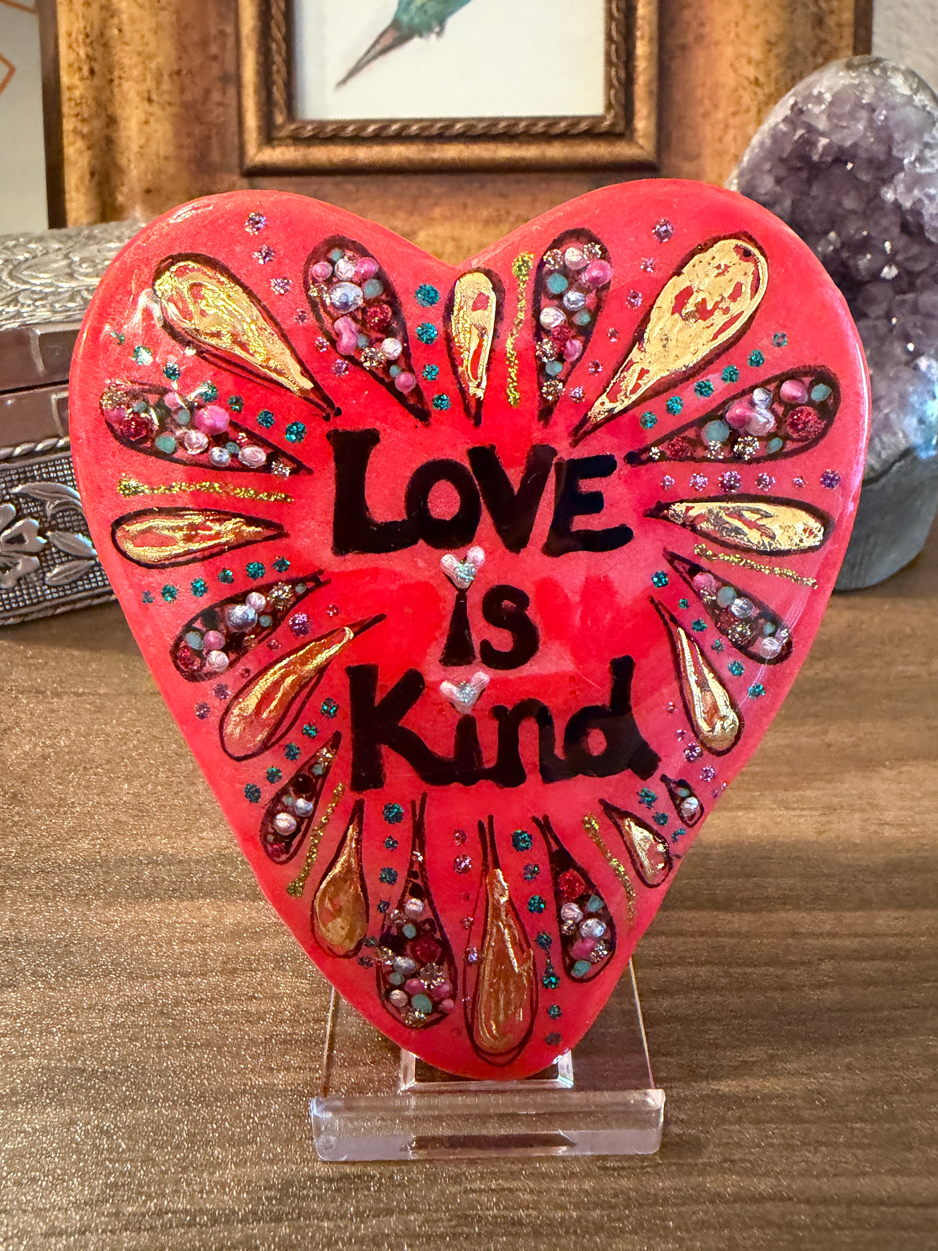 Decorative heart-shaped painted stone with 'Love is Kind' text on a stand.