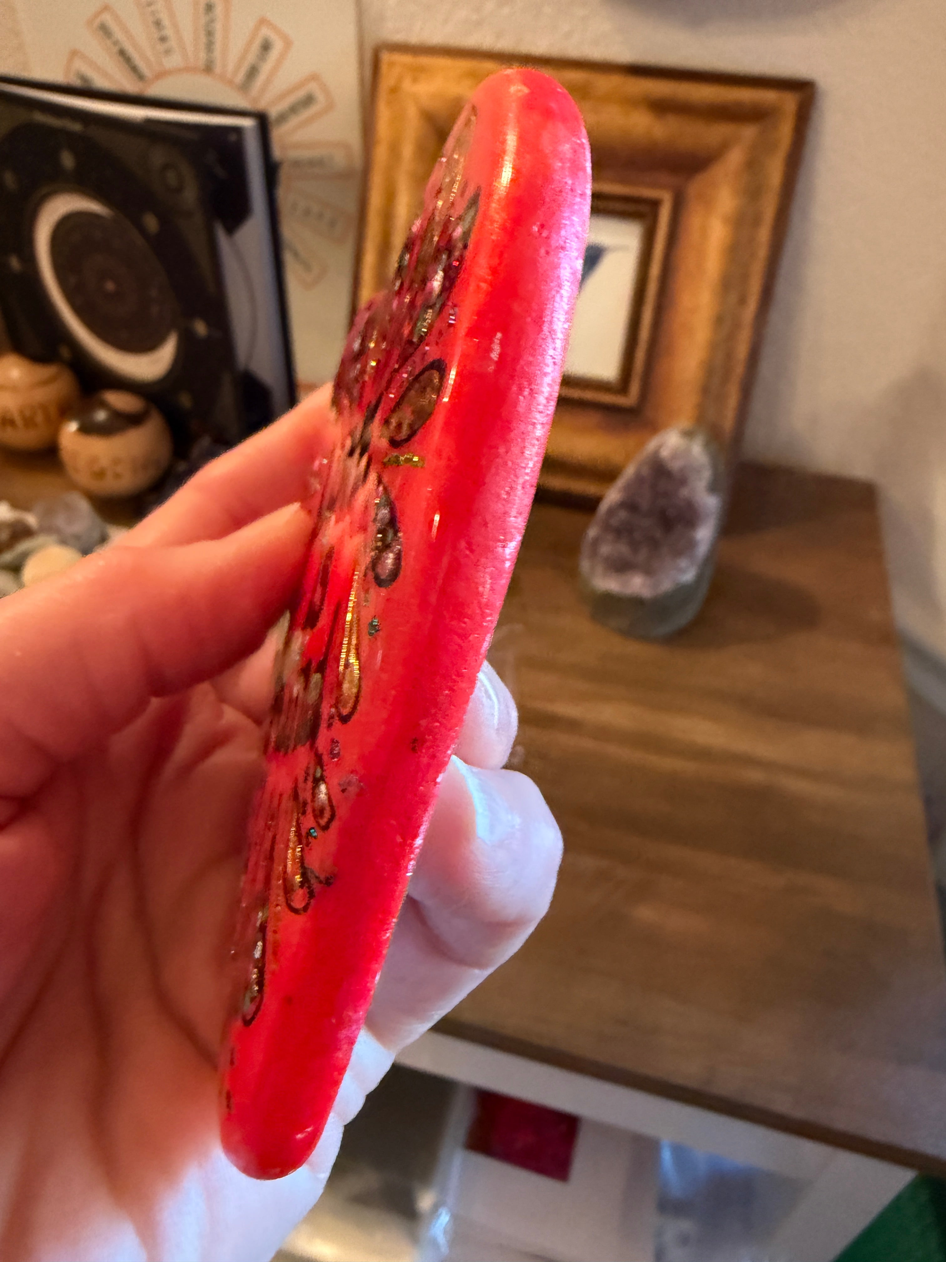 Hand holding a red painted stone with intricate designs in a casual indoor setting.