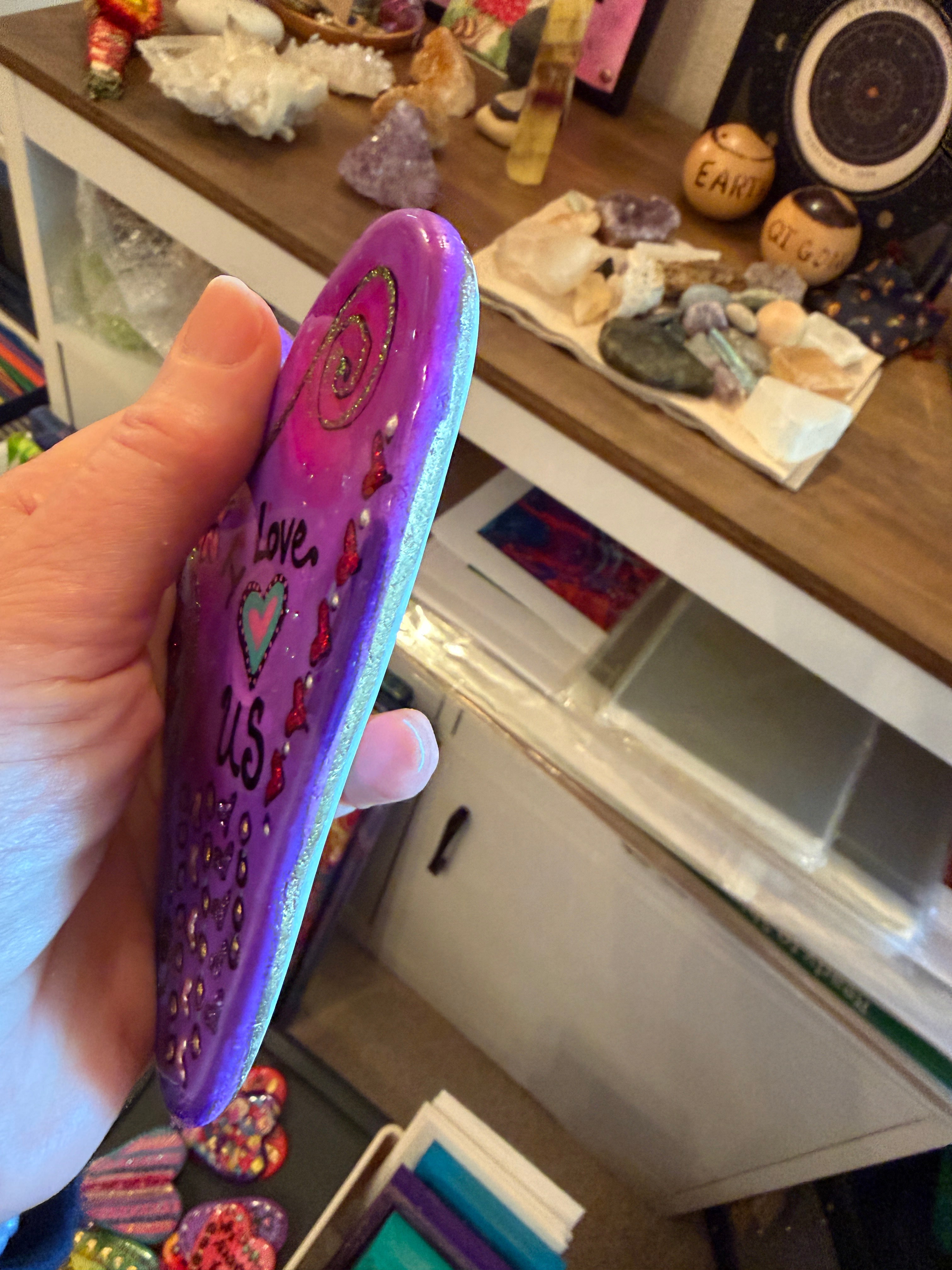 Hand holding a purple painted stone with decorative elements in a casual indoor setting