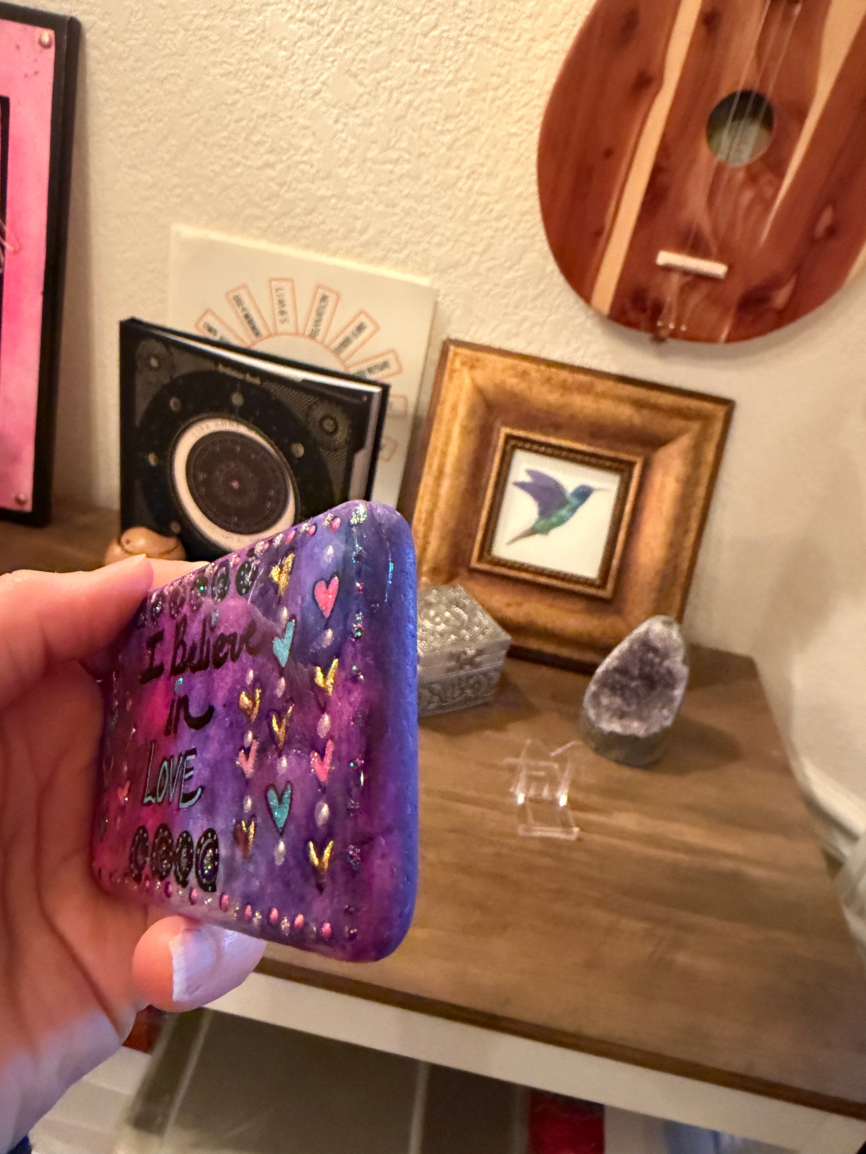Hand holding a  handpainted stone with designs in front of a wall with decorative items.