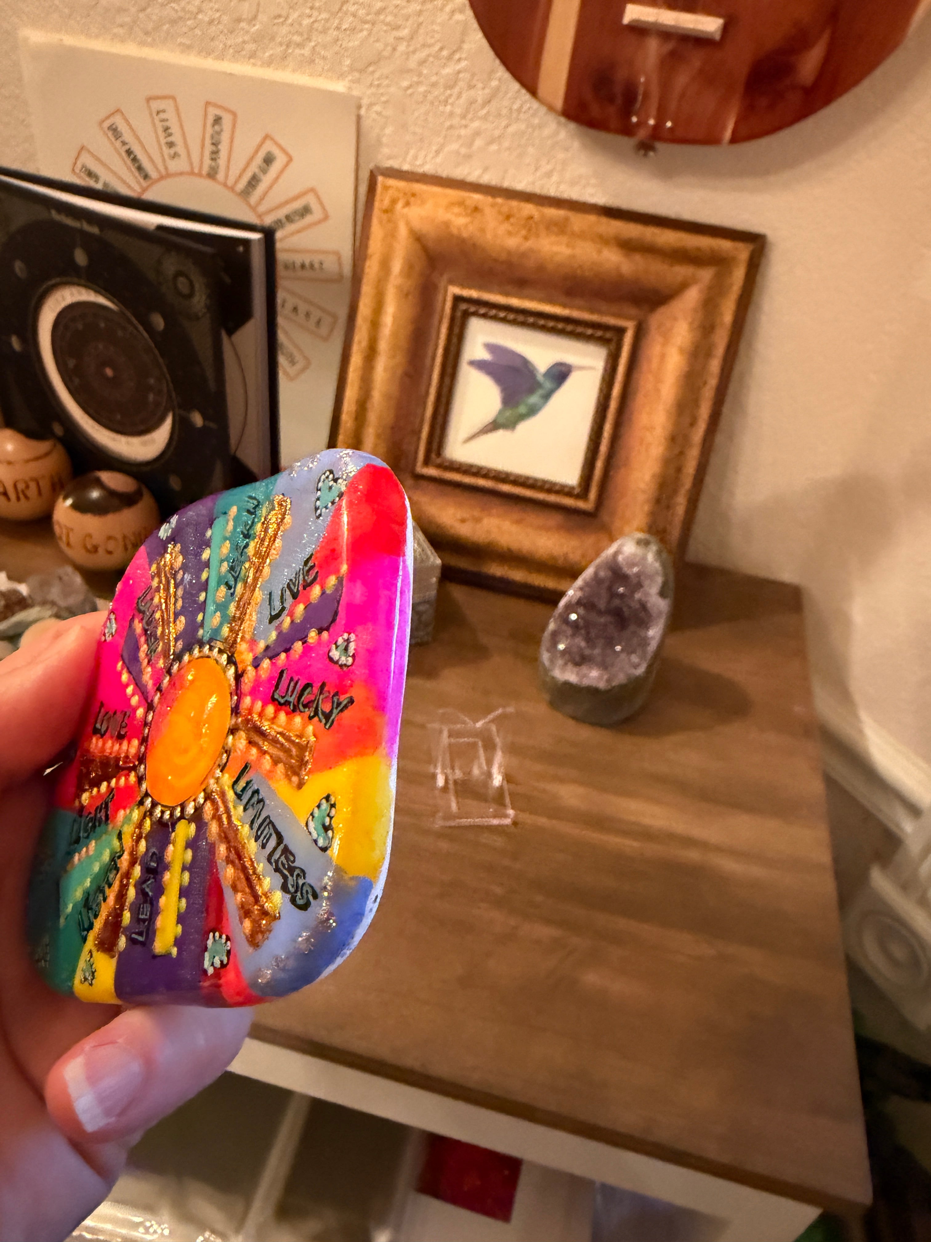 Colorful painted stone in front of a decorative setting with a framed picture and crystal on a wooden surface.