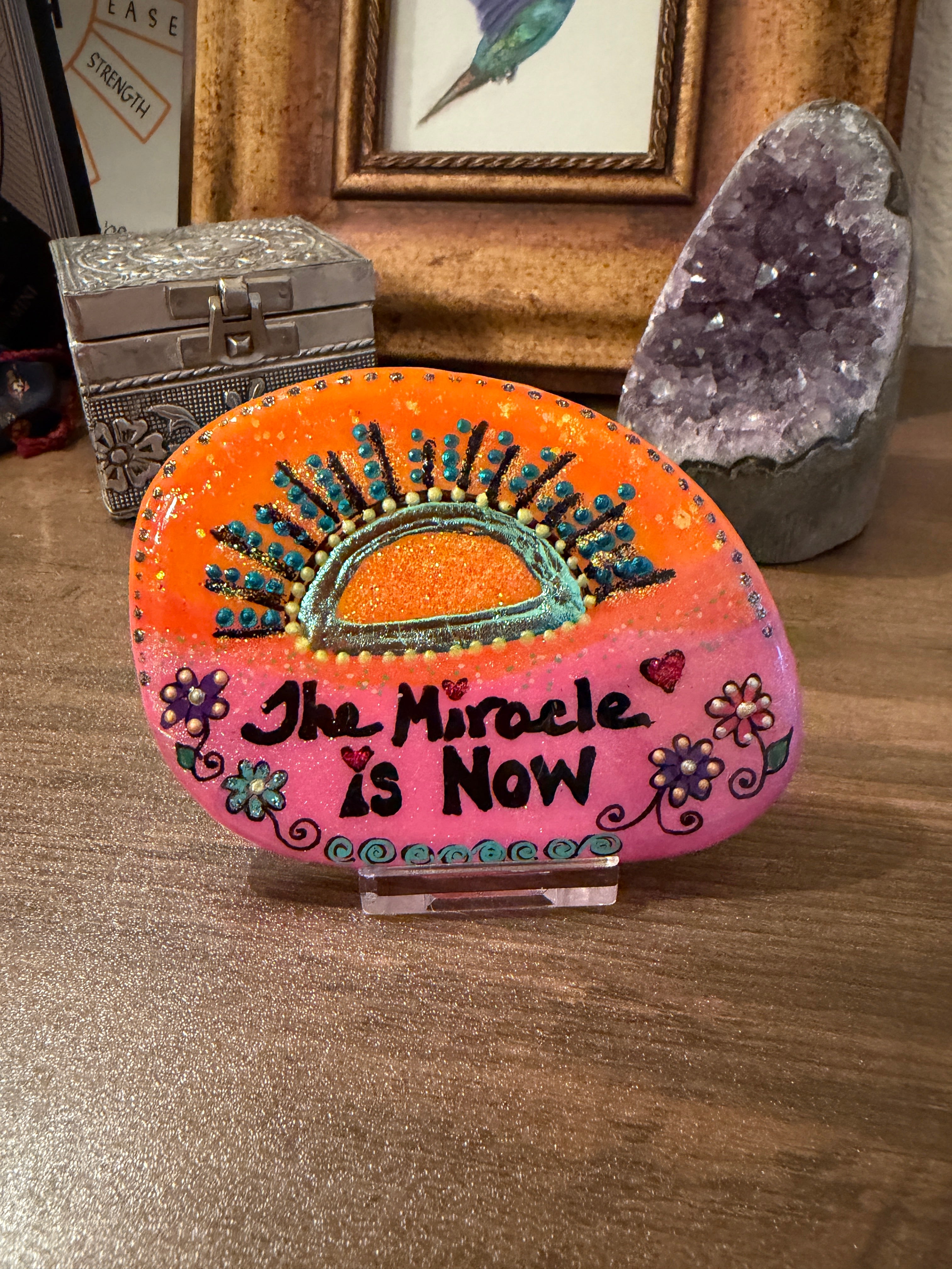 Decorative stone with 'The Miracle is Now' text on a wooden surface with crystals and a picture frame.