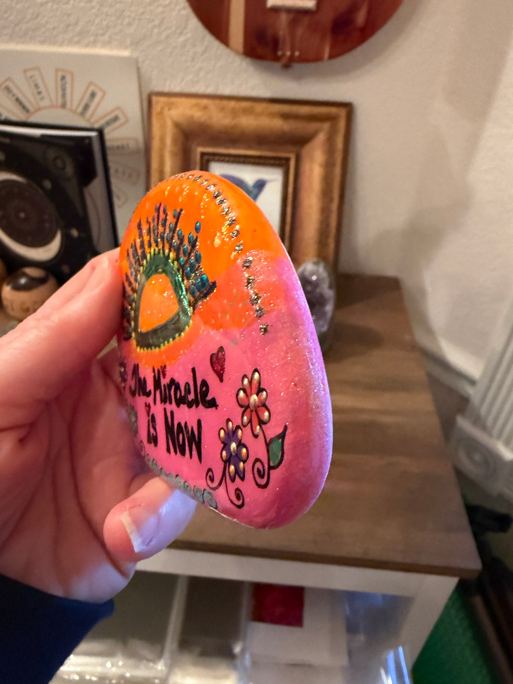 Hand holding a painted rock in pink and orange with an affirmation "the miracle is now"