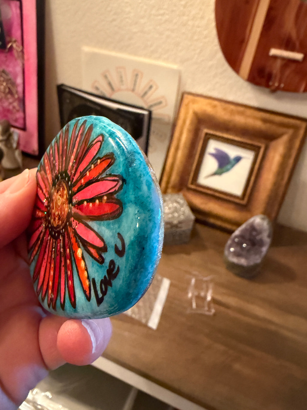 Hand holding a painted rock with a floral design in a room setting.