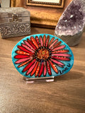 Decorative hand-painted stone with red and orange floral design on a blue background, placed on a wooden surface.